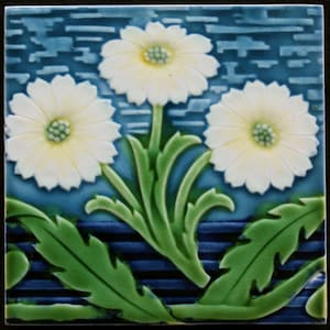 May include: A blue and white ceramic tile featuring a design of three white flowers with green leaves and stems. The background is a blue and white striped pattern.