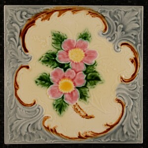 Può includere: Una piastrella di ceramica con un motivo floreale. La piastrella è decorata con due fiori rosa con centri gialli e foglie verdi. La piastrella è circondata da un bordo decorativo in tonalità di marrone e grigio.