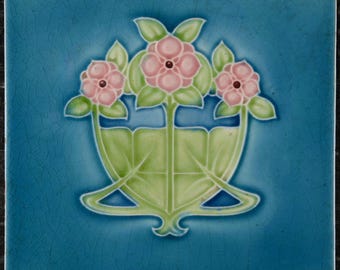 May include: Square ceramic tile with a teal blue background and a floral design. The design features three pink flowers with green leaves and stems, set in a green vase-like structure. The tile has a crackled texture.