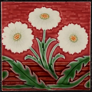 May include: Square ceramic tile with a design of three white daisy-like flowers with orange centres on a red background. Green leaves and stems are also visible. The tile has a decorative border.