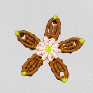 Könnte beinhalten: Eine braune und rosafarbene Makramee-Blume mit grünem Zentrum. Die Blume hat fünf Spitzen und ist mit einer Knotentechnik hergestellt.