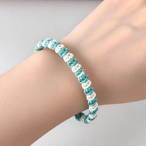 May include: A bracelet constructed from alternating turquoise and white cord, featuring a series of small, tightly tied knots arranged in a continuous, repeating pattern that encircles the wrist in a simple, uniform design.