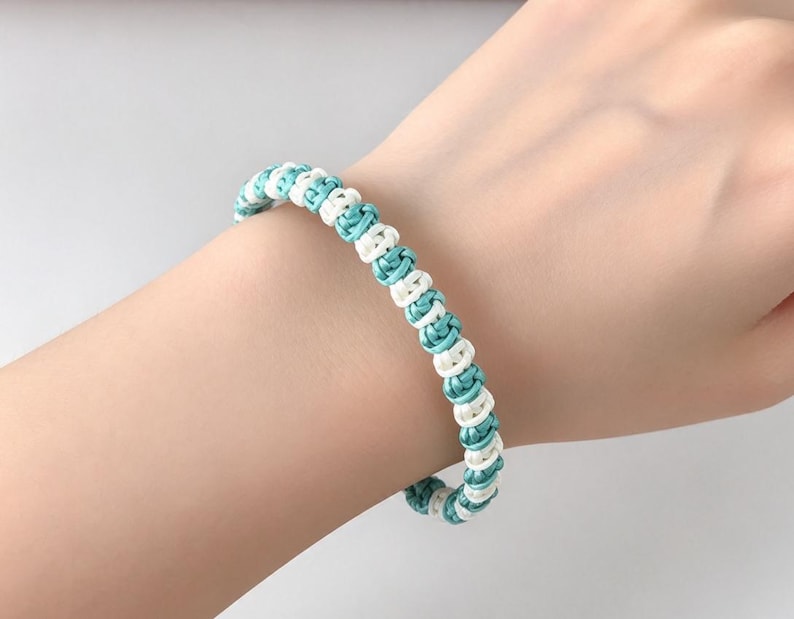 May include: A bracelet constructed from alternating turquoise and white cord, featuring a series of small, tightly tied knots arranged in a continuous, repeating pattern that encircles the wrist in a simple, uniform design.