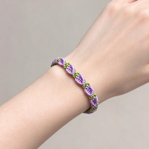 Op de afbeelding: Een handgemaakte macramé armband met een herhalend patroon van kleine, geknoopte paarse bloemachtige vormen verbonden door dunne, levendige limoengroene koordsegmenten, wat een delicaat en getextureerd geweven ontwerp creëert.