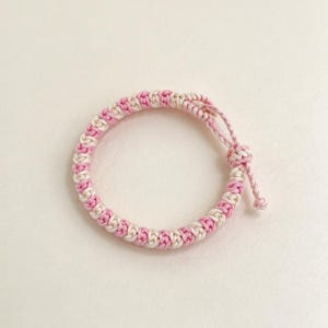 May include: A circular bracelet crafted from braided cord in alternating light pink and cream colors. The design features a repeating knotted pattern throughout the band, finished with a sliding knot closure and two short cord ends.