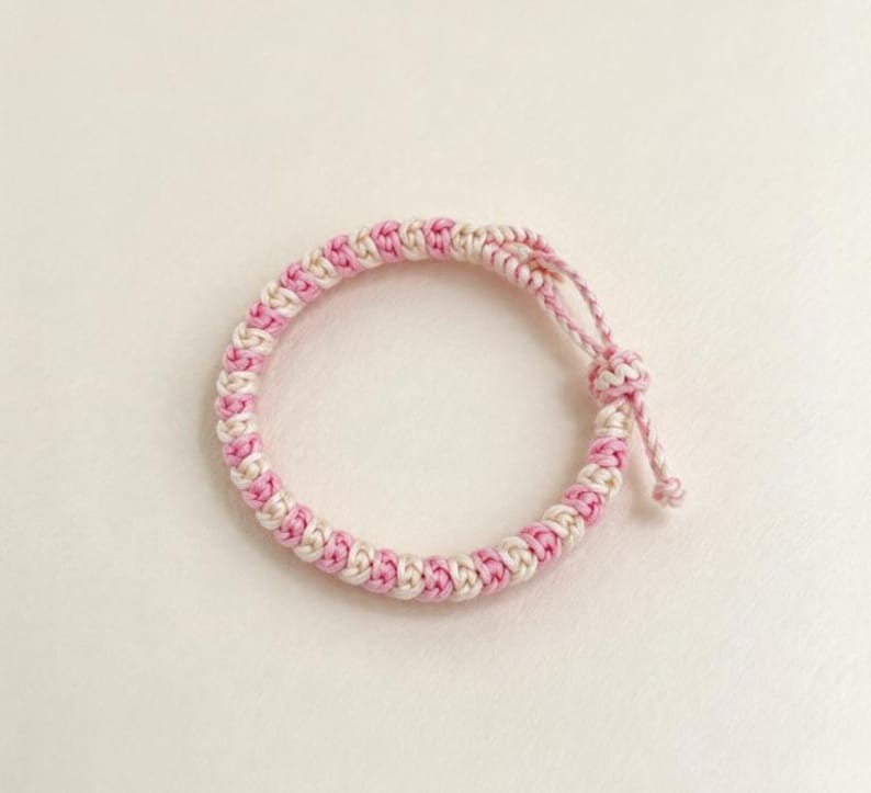 May include: A circular bracelet crafted from braided cord in alternating light pink and cream colors. The design features a repeating knotted pattern throughout the band, finished with a sliding knot closure and two short cord ends.