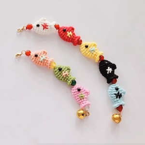 May include: Two crochet fish-shaped charms in various colors, including white, red, pink, green, yellow, black, and light blue. Each charm is connected with red beads and gold-colored clasps and bells. The charms are handmade.