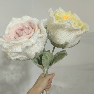 May include: A bouquet of two crocheted roses, one white with a pink centre and the other white with a yellow centre. The roses have green crocheted leaves.