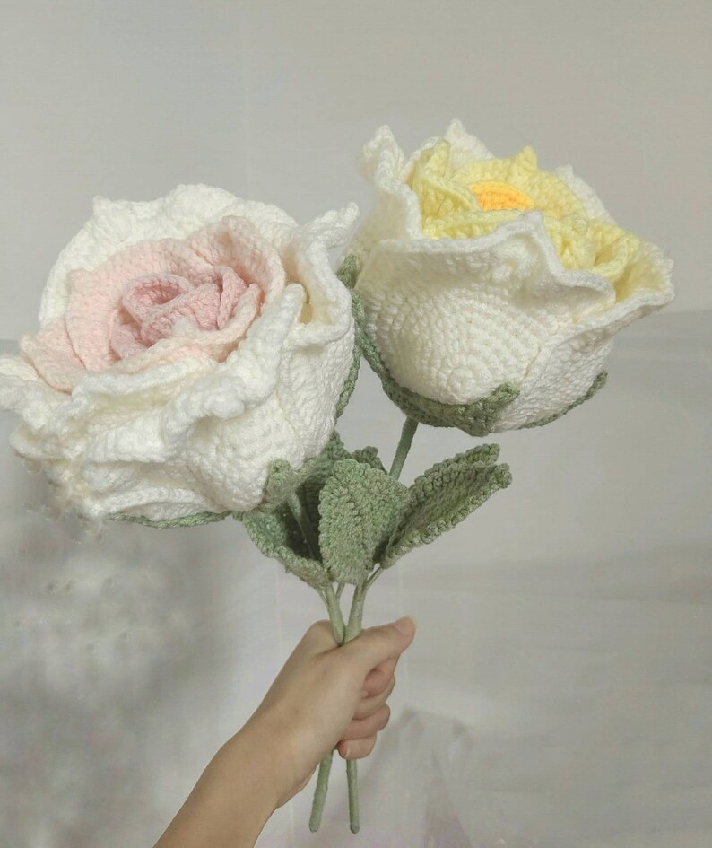 May include: A bouquet of two crocheted roses, one white with a pink centre and the other white with a yellow centre. The roses have green crocheted leaves.