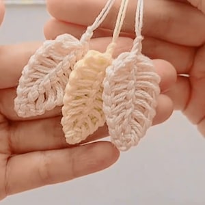 May include: Three crocheted leaf charms in white, cream, and light yellow. The charms are made with a delicate stitch pattern and have a slightly textured surface.
