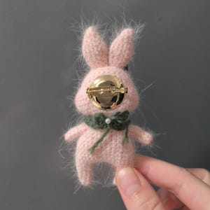 May include: A pink crocheted bunny brooch with a gold pin and a green scarf. The bunny has a fluffy texture and a pearl accent on the scarf.