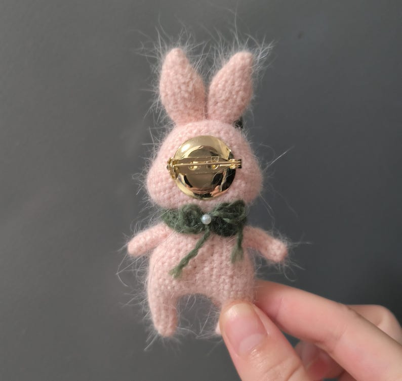 May include: A pink crocheted bunny brooch with a gold pin and a green scarf. The bunny has a fluffy texture and a pearl accent on the scarf.