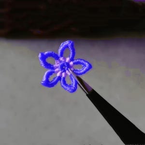 Pode incluir: Um close-up de um pequeno objeto azul em forma de flor segurado por pinças. A flor é feita de um material fino e flexível e tem um design delicado e intrincado.