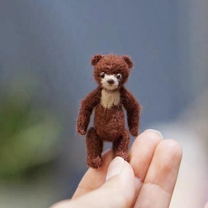 May include: A miniature brown teddy bear, approximately 2 inches tall, with a light tan chest and a detailed knitted texture. The bear has small, dark eyes and a slightly raised nose, giving it a lifelike appearance. The bear is a handmade item.