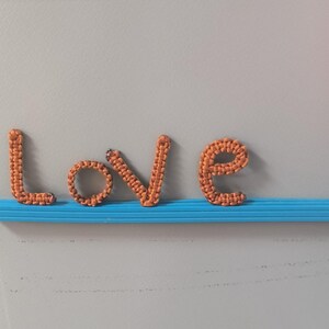 May include: Orange macrame letters spelling "LOVE" are displayed on a blue shelf against a gray background. The letters are crafted with a woven texture, creating a three-dimensional effect. The word is a decorative piece.