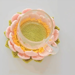 Może przedstawiać: Podkładka pod kubek w kształcie kwiatu lotosu wykonana na szydełku w odcieniach różowego, żółtego i zielonego. Podkładka jest przeznaczona do trzymania szklanki lub kubka.