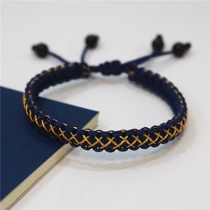 Peut inclure: Un bracelet tressé bleu marine et or avec une fermeture à nœud. Le bracelet est fait d'un cordon tissé avec un fil d'or qui le traverse.