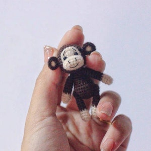 Puede incluir: Un peque&ntilde;o mono de crochet con pelaje marr&oacute;n, cara blanca y ojos negros. El mono est&aacute; siendo sostenido en una mano.