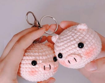 May include: Two handmade, light pink crochet pig keychains. Each pig has black button eyes, a stitched snout, and blush-colored cheeks. One keychain is attached to a silver keyring. The pigs are round and have small ears.
