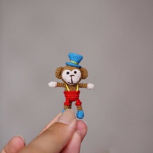 May include: A small crocheted monkey figurine featuring a brown body, white face, and round ears. The monkey wears a bright blue top hat with a yellow band, red shorts with yellow suspenders, and small blue shoes on its feet, all crafted in yarn.