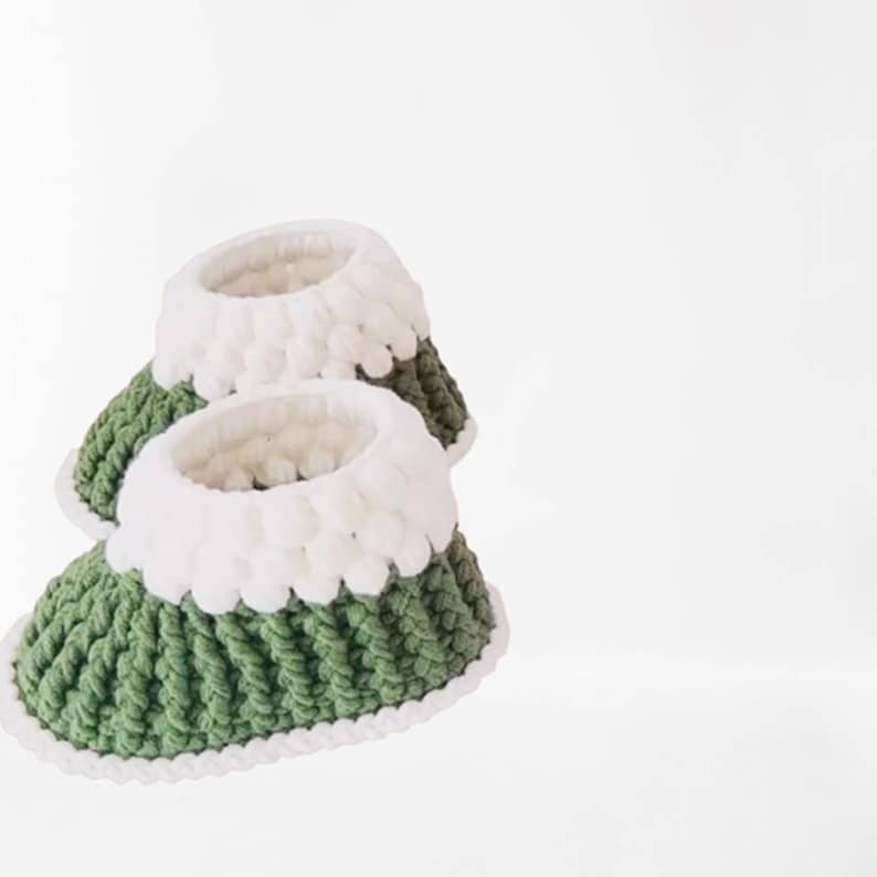 Puede incluir: Un par de botitas de beb&eacute; de crochet verde y blanco. Las botitas tienen un borde de crochet blanco alrededor de la parte superior y un cuerpo de crochet verde.