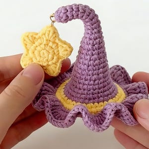 May include: Crocheted witch hat and star charm in shades of purple and yellow. The hat features a ruffled brim and a curved top, with a yellow band. The star charm is a bright yellow, adding a whimsical touch. The handmade item is held by two hands.