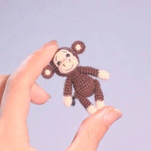 Puede incluir: Un peque&ntilde;o mono de crochet marr&oacute;n con una cara blanca y una expresi&oacute;n feliz. El mono est&aacute; siendo sostenido por una mano.