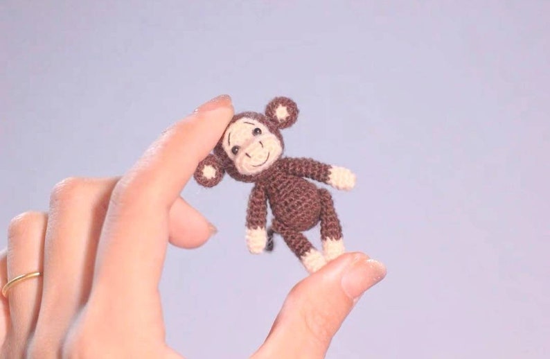 Puede incluir: Un peque&ntilde;o mono de crochet marr&oacute;n con una cara blanca y una expresi&oacute;n feliz. El mono est&aacute; siendo sostenido por una mano.