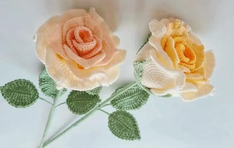 May include: Two crocheted roses with green leaves. One rose is peach and the other is yellow.