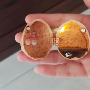May include: A hollowed-out walnut shell hinged to open like a locket, featuring a small internal LED light that emits a warm yellow glow. The interior contains a dark brown shelf and a small metallic battery component, with two tiny magnets for closure.