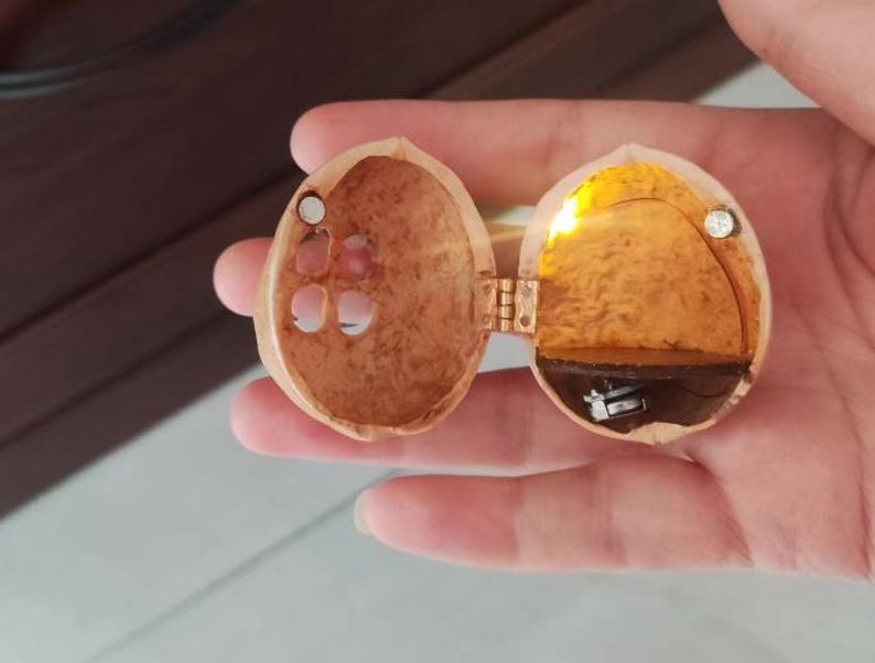 May include: A hollowed-out walnut shell hinged to open like a locket, featuring a small internal LED light that emits a warm yellow glow. The interior contains a dark brown shelf and a small metallic battery component, with two tiny magnets for closure.