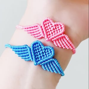 May include: Two macrame bracelets, one pink and one blue, each featuring a heart with wing details. The bracelets are handmade and appear to be crafted from a soft, woven material. The heart and wing design is a charming and unique accessory.