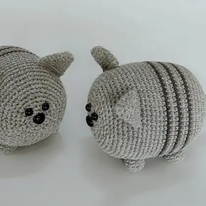 Pode incluir: Dois peluches de gato de crochê cinzentos com olhos pretos. Um gato tem um padrão listrado e uma cauda. O outro gato é de cor cinza sólida.