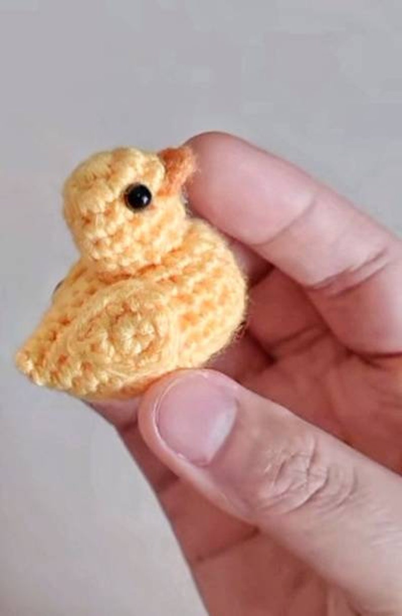 Puede incluir: Un peque&ntilde;o pollito amarillo tejido a crochet con ojos negros. El pollito est&aacute; siendo sostenido en una mano.