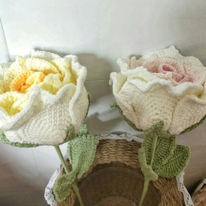 May include: Two crocheted roses, one in white and yellow, the other in white and pink, with green crocheted leaves. The roses are arranged in a vase.