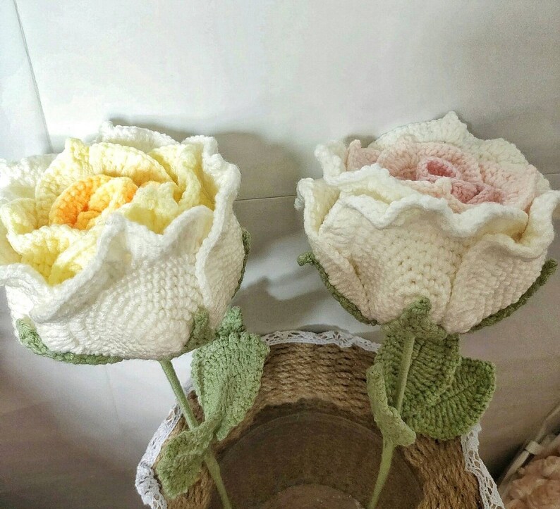 May include: Two crocheted roses, one in white and yellow, the other in white and pink, with green crocheted leaves. The roses are arranged in a vase.
