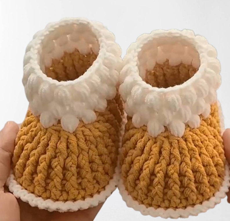 Puede incluir: Un par de botitas de beb&eacute; de crochet en amarillo y blanco. Las botitas tienen un borde de crochet blanco alrededor de la parte superior y un cuerpo de crochet amarillo.