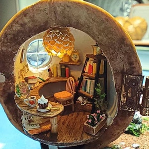 Pode incluir: Diorama em miniatura de um interior acolhedor, possivelmente uma casa de fadas, com uma janela redonda, uma secretária, uma estante e uma pequena mesa com uma chávena de café e um cupcake. O interior é iluminado por uma luz quente.