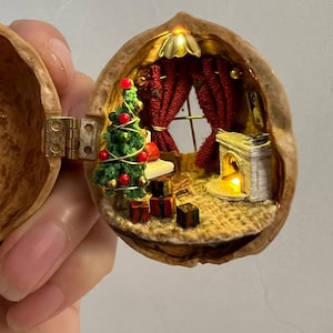 Pode incluir: Uma cena de Natal em miniatura dentro de uma casca de noz. A cena inclui uma pequena árvore de Natal, presentes, uma lareira e uma janela com cortinas vermelhas. O interior é iluminado com pequenas luzes, criando um brilho quente e festivo.