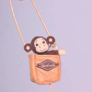 Puede incluir: Un peque&ntilde;o mono de crochet marr&oacute;n se sienta dentro de una bolsa de cuero marr&oacute;n con una correa de cadena dorada. La bolsa tiene una etiqueta de cuero marr&oacute;n que dice "Handmade".