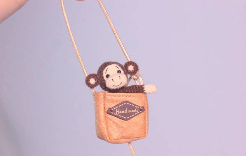 Puede incluir: Un peque&ntilde;o mono de crochet marr&oacute;n se sienta dentro de una bolsa de cuero marr&oacute;n con una correa de cadena dorada. La bolsa tiene una etiqueta de cuero marr&oacute;n que dice "Handmade".
