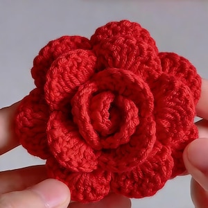 May include: A handmade, vibrant red crochet flower. The flower is in full bloom, with multiple layers of petals creating a realistic rose shape. The texture of the yarn is visible, adding depth and detail to the floral design. A craft item.