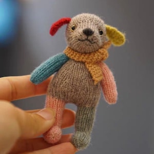 May include: A small knitted toy dog with a gray body, blue, pink, and yellow legs, and red and yellow ears. The dog is wearing a scarf in orange and yellow.