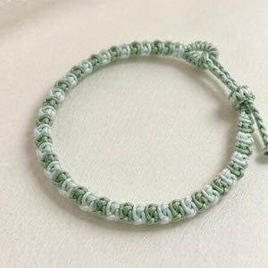 May include: A braided cord bracelet featuring alternating sage green and pale mint green threads knotted in a repeating pattern. The bracelet includes a sliding knot closure with two short cord ends, each finished with a small decorative knot.