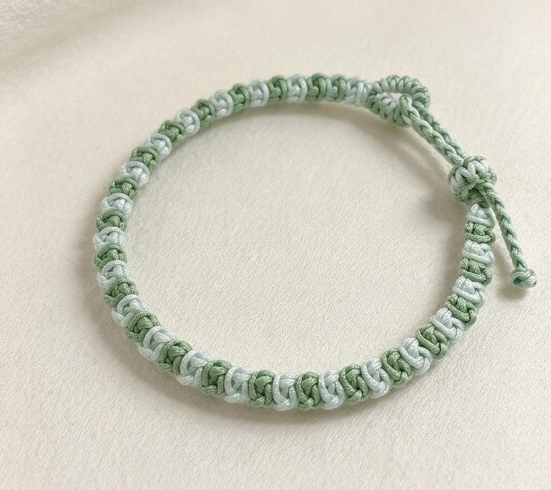May include: A braided cord bracelet featuring alternating sage green and pale mint green threads knotted in a repeating pattern. The bracelet includes a sliding knot closure with two short cord ends, each finished with a small decorative knot.