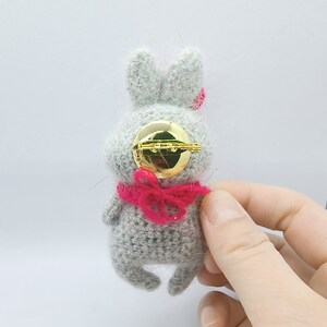 May include: A gray crocheted bunny brooch with a pink bow and a gold pin back.