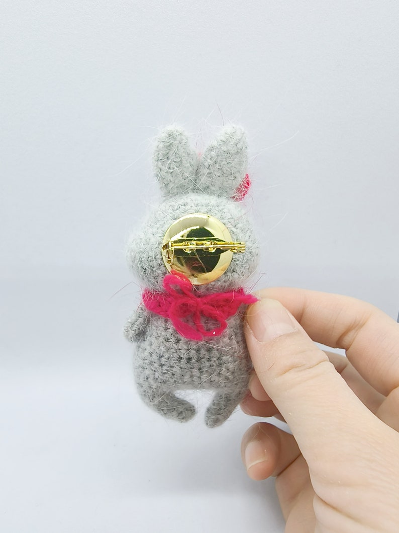 May include: A gray crocheted bunny brooch with a pink bow and a gold pin back.