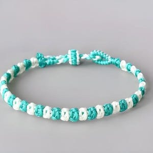 May include: A woven cord bracelet featuring an alternating pattern of bright turquoise and white thread. The design consists of a series of small, uniform knots arranged in a continuous circular band with a sliding knot closure mechanism.