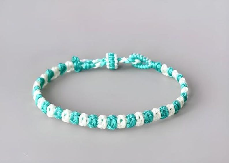 May include: A woven cord bracelet featuring an alternating pattern of bright turquoise and white thread. The design consists of a series of small, uniform knots arranged in a continuous circular band with a sliding knot closure mechanism.