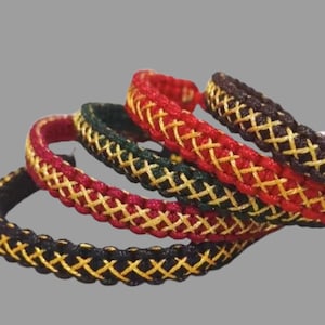 K&ouml;nnte beinhalten: Vier geflochtene Armb&auml;nder mit einem Kreuzmuster. Die Armb&auml;nder sind schwarz, rot, gr&uuml;n und braun mit goldfarbenem Garn, das in das Design eingewebt ist.